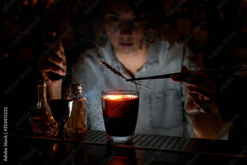 Woman barman decorates red cocktail with spikelet using tweezers and sprinkles on it.