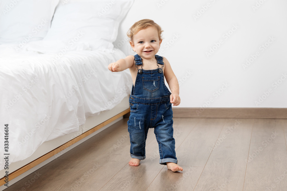 Cute baby in overalls making first steps in bedroom Cute baby in overalls making first steps in bedroom