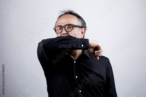 uomo in camicia nera con occhiali starnutisce e si ripara con il gomito, isolato su sfondo nero photo