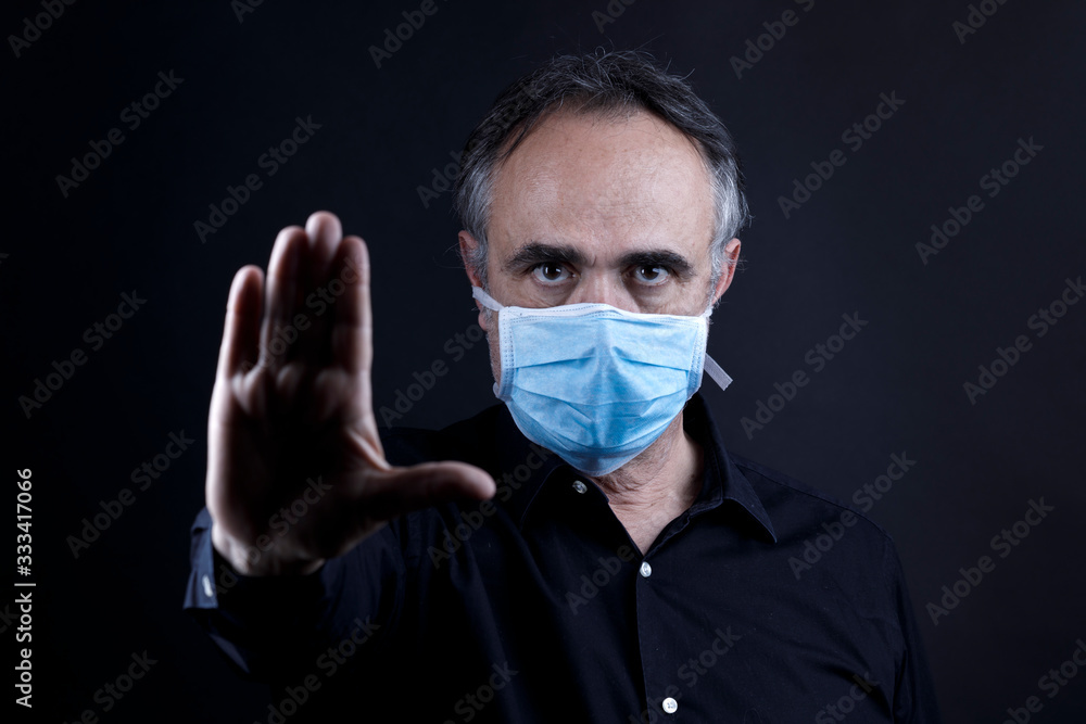 uomo in camicia nera che indossa una mascherina azzurra, isolato su sfondo nero fa cenno di stop con la mano uomo in camicia nera che indossa una mascherina azzurra, isolato su sfondo nero fa cenno di stop con la mano