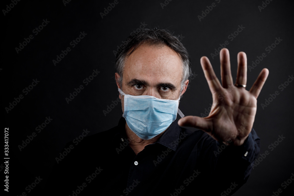 uomo in camicia nera che indossa una mascherina azzurra, isolato su sfondo nero fa cenno di stop con la mano uomo in camicia nera che indossa una mascherina azzurra, isolato su sfondo nero fa cenno di stop con la mano