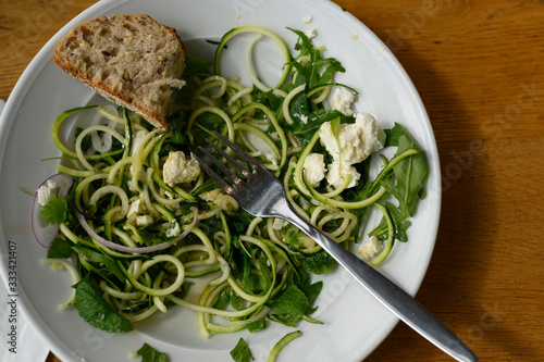 Unfinished salad on a wooden table. A cold appetizer of zucchini noodles, with fresh herbs and feta.On a white plate lies a piece of bread and a fork.Vegetarian version of the dish. Healthy nutrition.