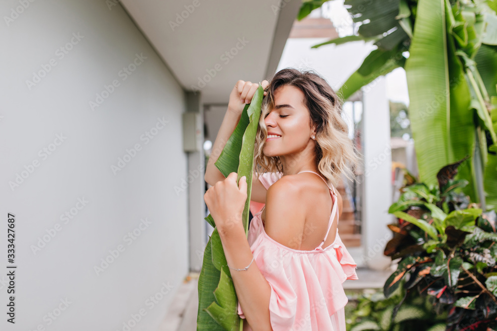 Amazing girl with cute haircut posing with eyes closed near garden. Outdoor photo of ecstatic tanned woman in trendy pink dress. Amazing girl with cute haircut posing with eyes closed near garden. Outdoor photo of ecstatic tanned woman in trendy pink dress.