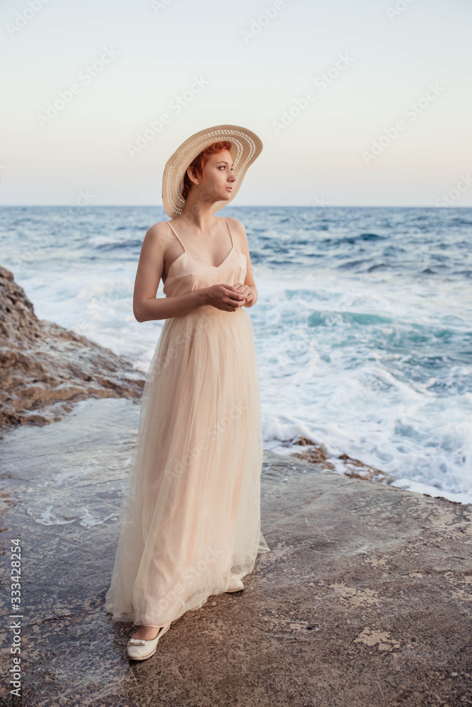A beautiful, red-haired girl is standing against the sea, wearing a hat on her head, wearing an airy, light dress. Waiting for a lover, the heroine of the novel. A beautiful, red-haired girl is standing against the sea, wearing a hat on her head, wearing an airy, light dress. Waiting for a lover, the heroine of the novel.