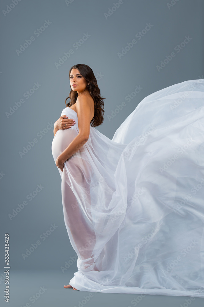 Studio portrait of nude pregnant woman in a white flying dress fabric on grey background. Beautiful healthy pregnancy, happy motherhood Studio portrait of nude pregnant woman in a white flying dress fabric on grey background. Beautiful healthy pregnancy, happy motherhood
