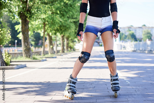 Young woman, roller-blading park with green trees in summer morning at sunny weather. Tan legs, wearing roller-skates and protective equipment, showing the process of riding in town.