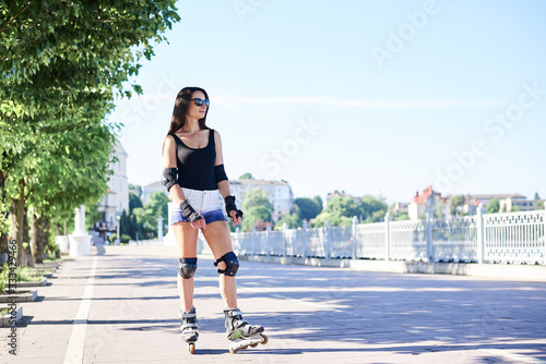 Young brunette woman with long hair, wearing rollerblades, posing in city park in the morning. Standing full-length portrait of roller-skater, wearing black top and white shorts. Summer activities.