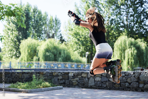 Young brunette athlete, wearing rollerblades and protective outfit, jumping in the air while riding in green city park in summer morning. Slim fit woman, riding roller-skates, training.