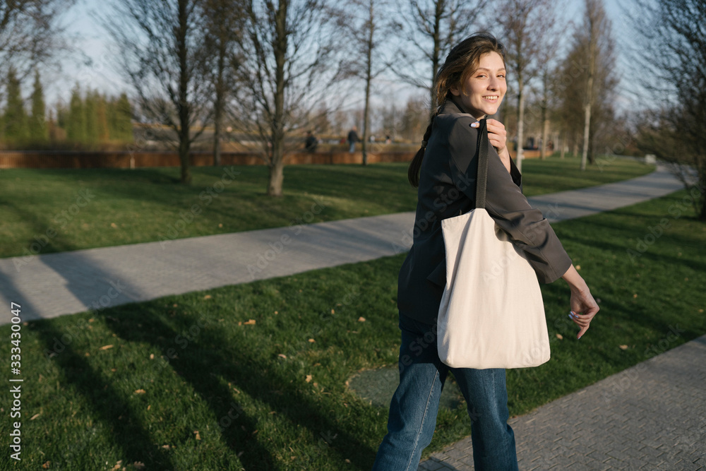 Beautiful smiling woman with an eco white bag on her shoulder walks in the park and looks at the camera. Millennials go shopping Beautiful smiling woman with an eco white bag on her shoulder walks in the park and looks at the camera. Millennials go shopping