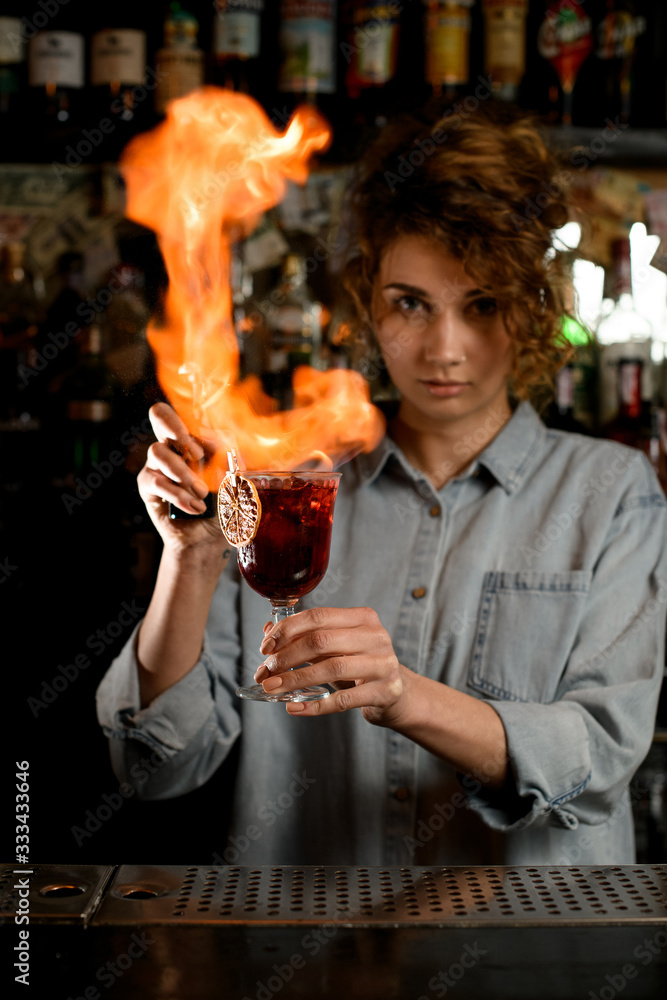 Professional woman barman holds wineglass with cocktail and makes flame over it.