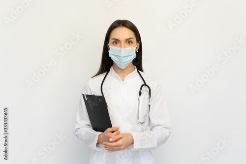 Friendly female doctor in a protective medical mask with a stethoscope, holds a folder in his hands,