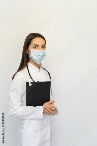 Friendly female doctor in a protective medical mask with a stethoscope, holds a folder in his hands,