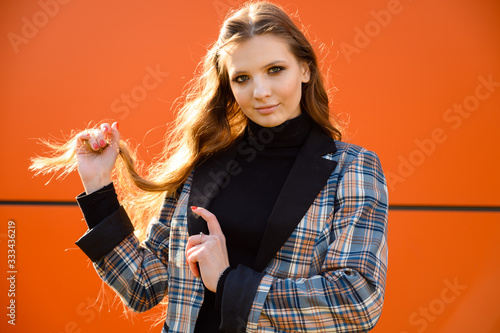 Portrait of a stylish beautiful young girl with long hair with a smile in the park against the background of an orange building wall. Photo taken in sunny weather outdoors in spring.