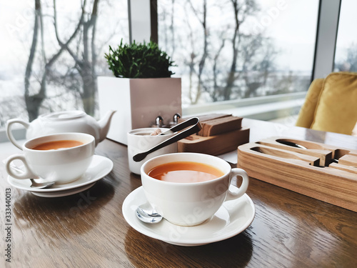 Two cups of healthy herbal tea with sea-buckthorn, ginger and honey on a textured light wooden table in a cafe with modern trendy interior. Mockup for your design.