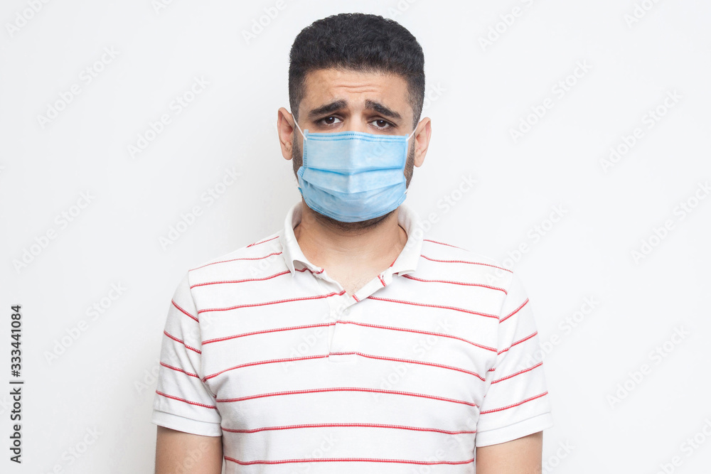 Portrait of sad upset or bored man with surgical medical mask in striped t-shirt standing and looking at camera with dissatisfied sadness face. indoor studio shot, isolated on white background. Portrait of sad upset or bored man with surgical medical mask in striped t-shirt standing and looking at camera with dissatisfied sadness face. indoor studio shot, isolated on white background.