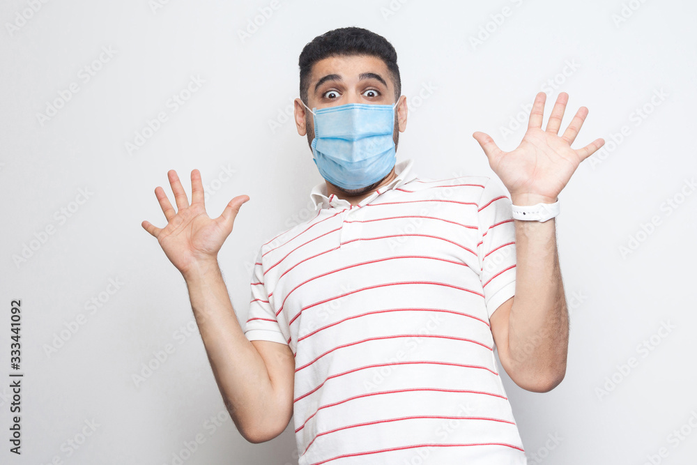 Portrait of shocked young with surgical medical mask man in striped t-shirt standing with raised arms and looking at camera with big eyes and panic. indoor studio shot, isolated on white background. Portrait of shocked young with surgical medical mask man in striped t-shirt standing with raised arms and looking at camera with big eyes and panic. indoor studio shot, isolated on white background.