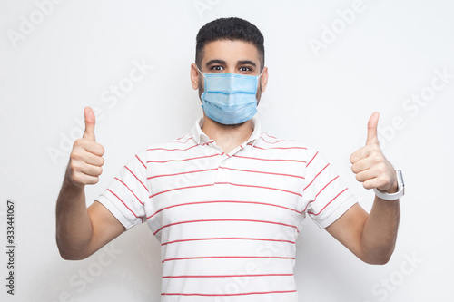 I like this. I am safe. Portrait of satisfied happy young man with surgical medical mask in striped t-shirt standing, thumbs up and looking at camera. indoor studio shot, isolated on white background.