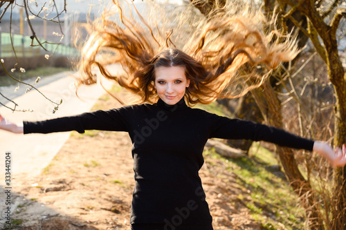 Portrait of a pretty young girl playing with long hair with a smile on the background of road trees in the park. Photo in sunny weather outdoors in spring.