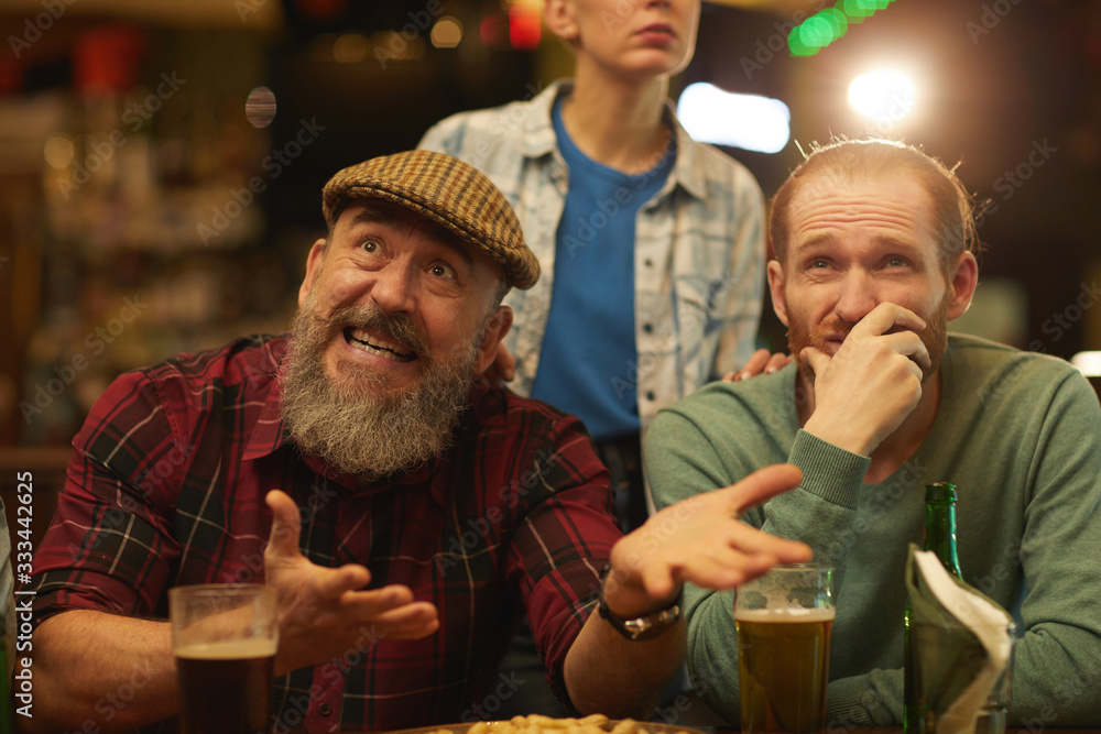 Mature men gesturing and looking unsatisfied while watching football match and drinking beer in sport bar