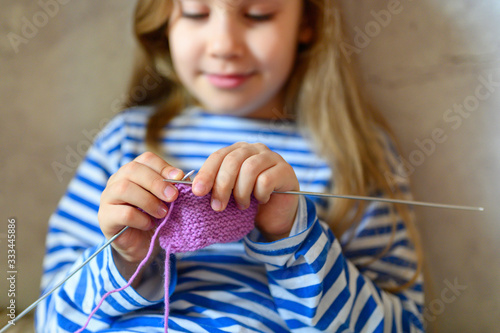 kid girl knitting with needles in home interior on background of gray concrete wall. the concept of teaching and studies at home during the quarantine
