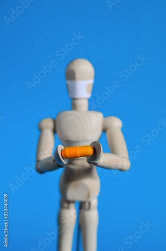 A wooden figure in a medical mask holds an orange pill on a blue background. Coronavirus  pandemic and epidemic concept