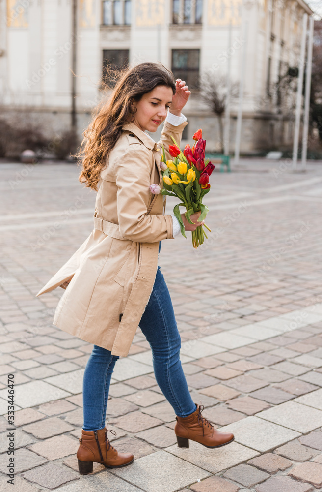 Fototapeta premium Beautiful young woman with spring tulips flowers bouquet at city street. Happy girl walking outdoors. Spring portrait of pretty female in old town