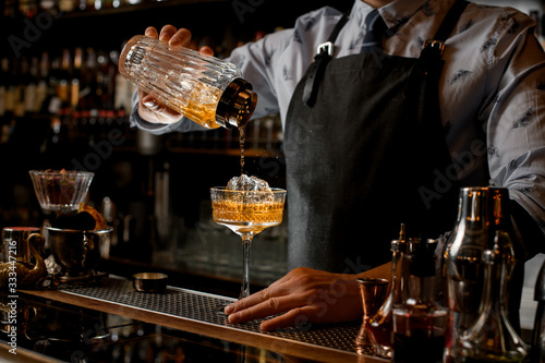 barman regularly pours finished cold cocktail from glass shaker into wineglass.