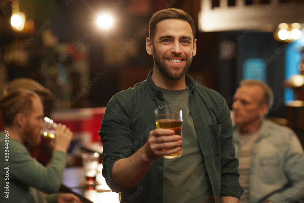 Portrait of young man in casual clothing smiling at camera and drinking beer with his friends in the pub