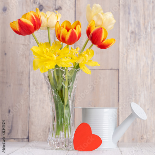 Tulip flower arrangement in glass vase with heart greeting, watering can decor on wooden table background wall, close up, Mother's Day design concept.