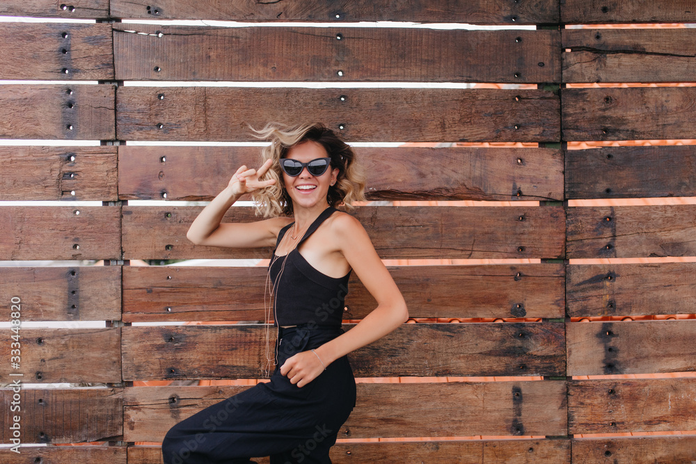 Inspired tanned woman expressing energy while posing outdoor. Photo of laughing young lady in black clothes isolated on wooden background. Inspired tanned woman expressing energy while posing outdoor. Photo of laughing young lady in black clothes isolated on wooden background.