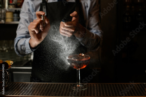 Bartender in black apron sprinkles on glass with cocktail.