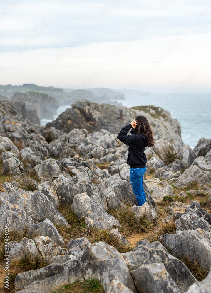 Beautiful young girl with black sweater standing on rocks beside cliffs at the coast in a beautiful landscape
