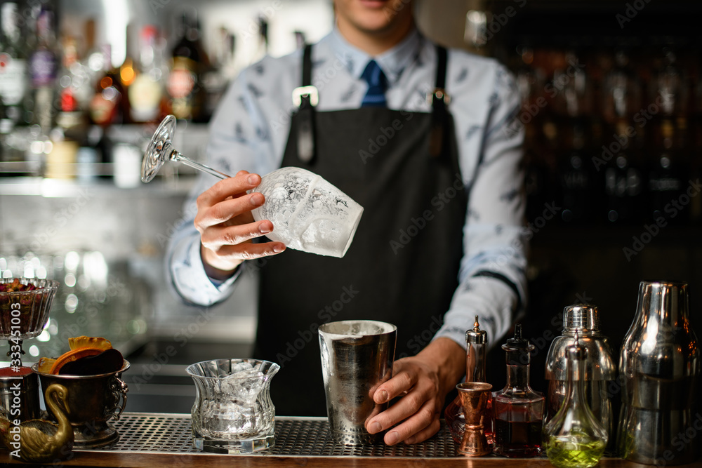 bartender in black apron holds glass and steel shaker at bar.