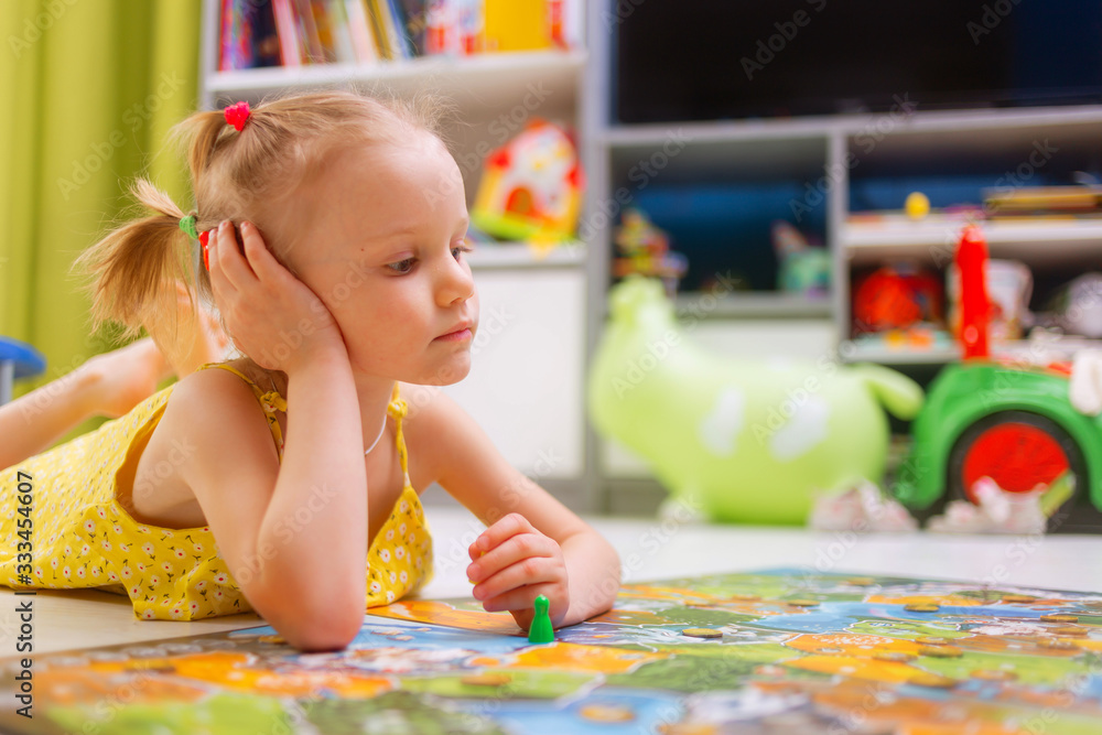 Fototapeta premium Board game concept. Little kid girl play in board game at home sitting on a wooden floor. Board game field, many figures.