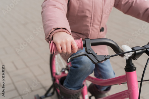The child is holding the wheel of a bicycle. A girl in a pink jacket is riding her bike.