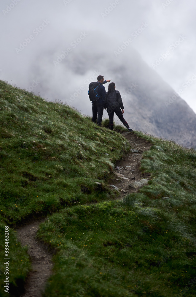 Back view of young couple standing on grassy hill and admiring the view of majestic foggy mountain. Male traveler pointing at something while holding girlfriend hand. Concept of hiking, relationships.