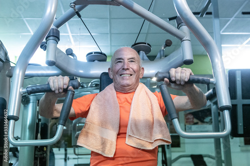 Uomo anziano con maglia arancione e asciugamano al collo si allena in palestra nei macchinari, 