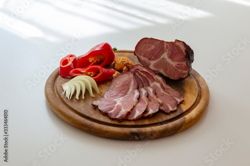 Sliced smoked pork neck with red pepper, garlic cloves and onion slices on a wooden cutting board.
