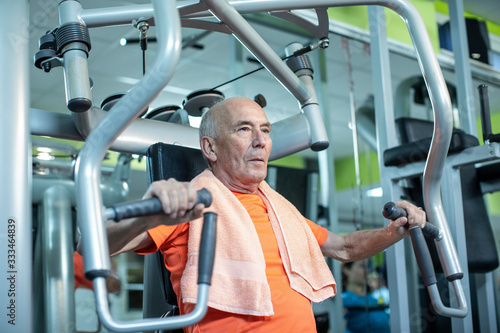 Uomo anziano con maglia arancione e asciugamano al collo si allena in palestra nei macchinari