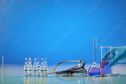 Coronavirus. Hand in glove holding beaker with purple reagent. Six ampoules with liquid, syringe, medical flask and phonendoscope, blue background.