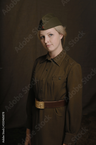 Portrait young blonde woman in military uniform from second world war stands and looks at lens of camera on brown background