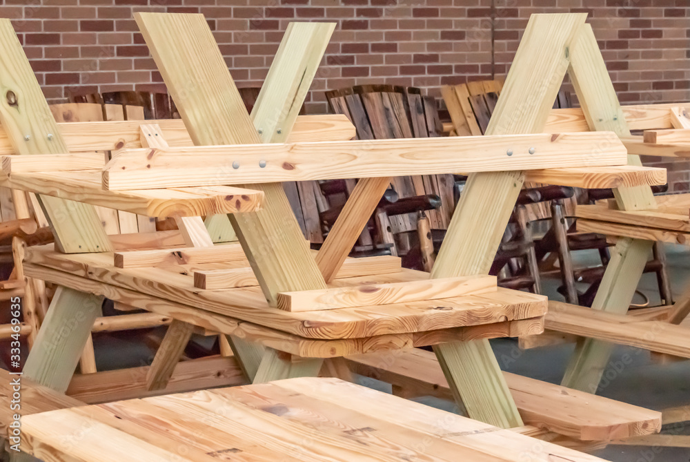 Stacked unfinished wooden picnic tables and rocking chairs for sale outside against a brick wall.

