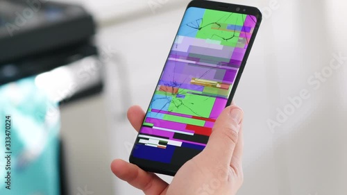 A close up on a hand holding a smart mobile phone devise with a broken screen  with cracks and a colorful blue, green, pink lost signal on damaged display,