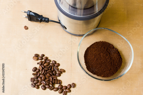 Coffee grinding at home. Electric metal coffee grinder, roasted coffee beans and ground coffee in a glass bowl on a beige table. The view from the top