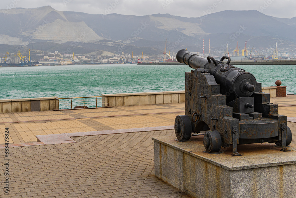 Novorossiysk, Russia - March 25, 2020. - Embankment of Admiral Serebryakov. Old naval gun is directed towards the sea. Coast of Tsemess bay. Blurred background of port cranes at terminals of seaport. Novorossiysk, Russia - March 25, 2020. - Embankment of Admiral Serebryakov. Old naval gun is directed towards the sea. Coast of Tsemess bay. Blurred background of port cranes at terminals of seaport.
