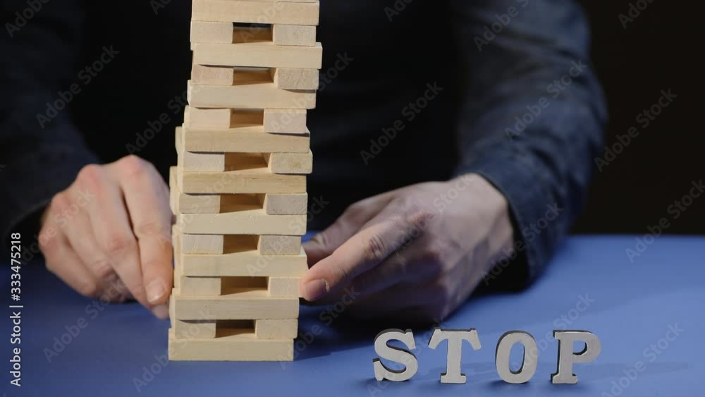 Male hand pulls out a wooden block and tower collapses destroying the word stop on the table. Risks in the business. Jenga falls on the table
