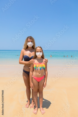 A woman and a child in a protective surgical mask on their face are standing on the beach in swimsuits. Chinese coronavirus disease COVID-19 is dangerous