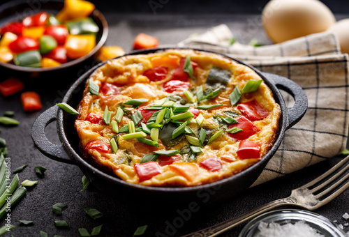 Frittata with the addition of multi-colored peppers and green onions in a cast-iron baking dish on a black background close up