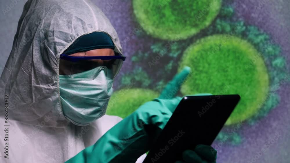 A worker in a hazmat suit with protective shield glasses, a face mask and gloves working on his tablet computer device on a virus bacteria microscopic view background.Public Domain background element A worker in a hazmat suit with protective shield glasses, a face mask and gloves working on his tablet computer device on a virus bacteria microscopic view background.Public Domain background element