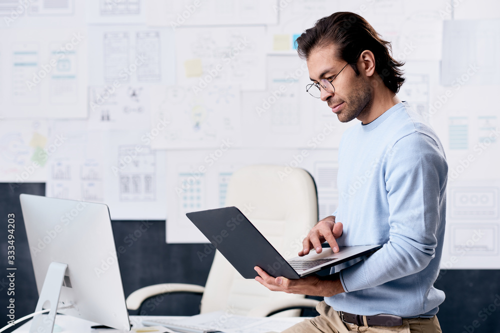 Serious modern middle-aged man in glasses using laptop while analyzing interfaces of web sites in UI design office Serious modern middle-aged man in glasses using laptop while analyzing interfaces of web sites in UI design office